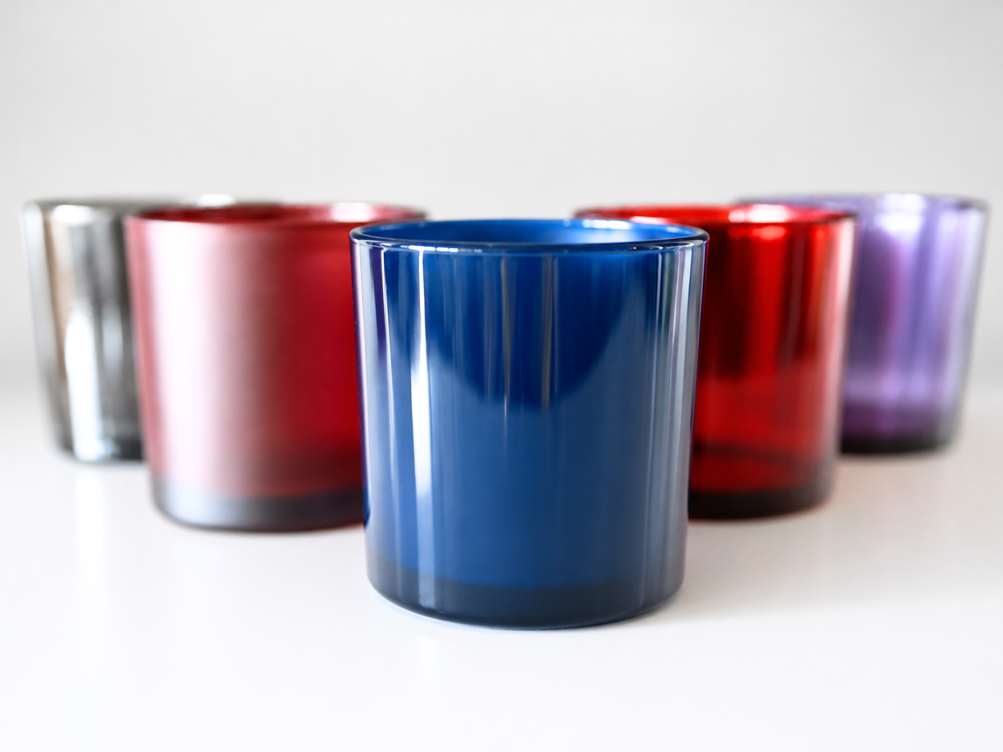 A set of six translucent glass candle holders in various colors, including blue, red, purple, pink, silver, and dark blue, arranged in a row on a white surface with a light gray background.