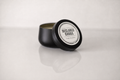 A black metal tin candle with its lid leaning against the container, showing a white circular label that reads 'DECLARED GOODS'. The candle is set on a reflective white surface against a light gray background.