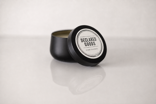 A black metal tin candle with its lid leaning against the container, showing a white circular label that reads 'DECLARED GOODS'. The candle is set on a reflective white surface against a light gray background.