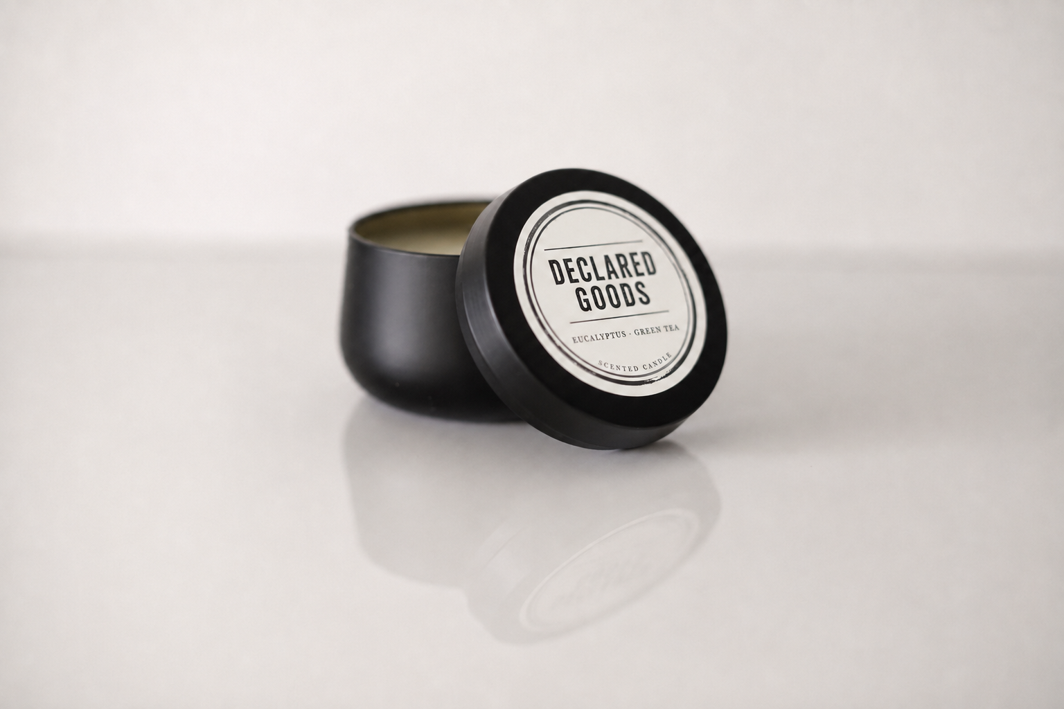 A black metal candle tin with its lid leaning against it, showing a white circular label that reads 'DECLARED GOODS' and 'PEPPERMINT + SMOKED SUGAR OIL' on a light gray background.