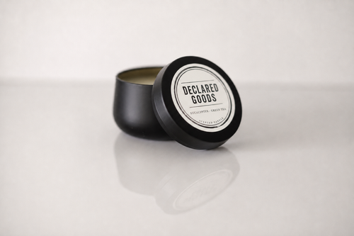 A black metal candle tin with its lid leaning against it, showing a white circular label that reads 'DECLARED GOODS' and 'PEPPERMINT + SMOKED SUGAR OIL' on a light gray background.
