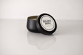 A black metal tin candle with its lid leaning against the container, showing a white circular label that reads 'DECLARED GOODS'. The candle is set on a reflective white surface against a light gray background.