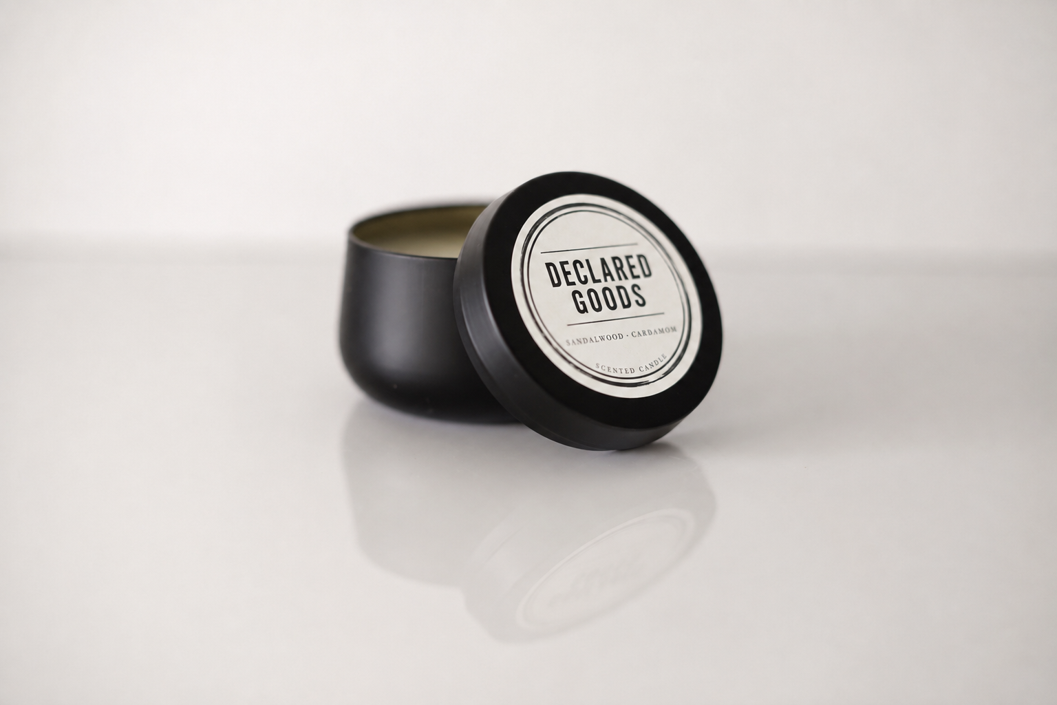 A black metal tin candle with its lid leaning against the container, showing a white circular label that reads 'DECLARED GOODS'. The candle is set on a reflective white surface against a light gray background.