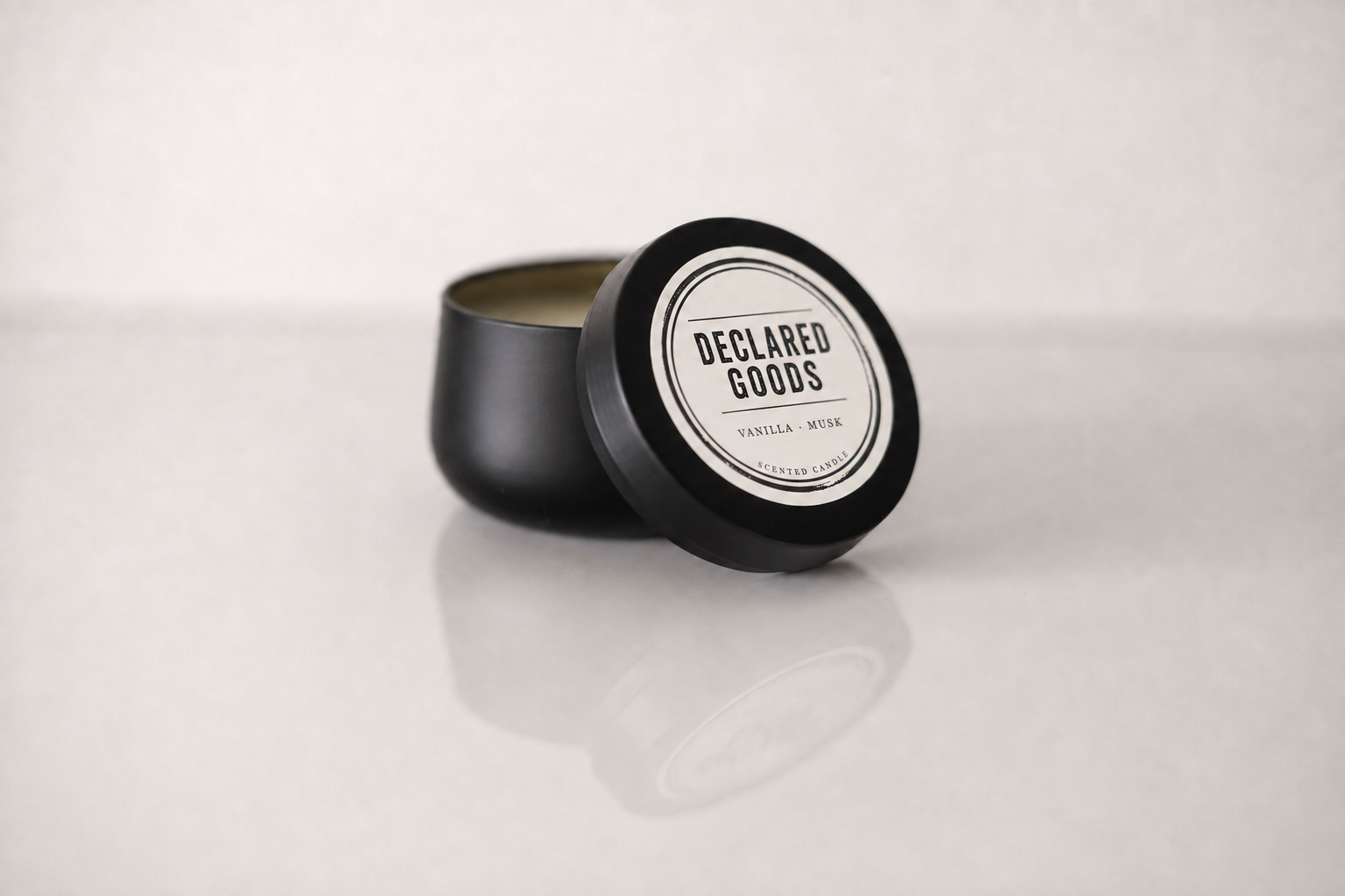 A black metal candle tin with its lid leaning against it, showing a white circular label that reads 'DECLARED GOODS' and 'PEPPERMINT + SMOKED SUGAR OIL' on a light gray background.